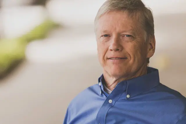 An older man in a blue dress shirt sits comfortably outdoors, with a blurred background of greenery and soft lighting.