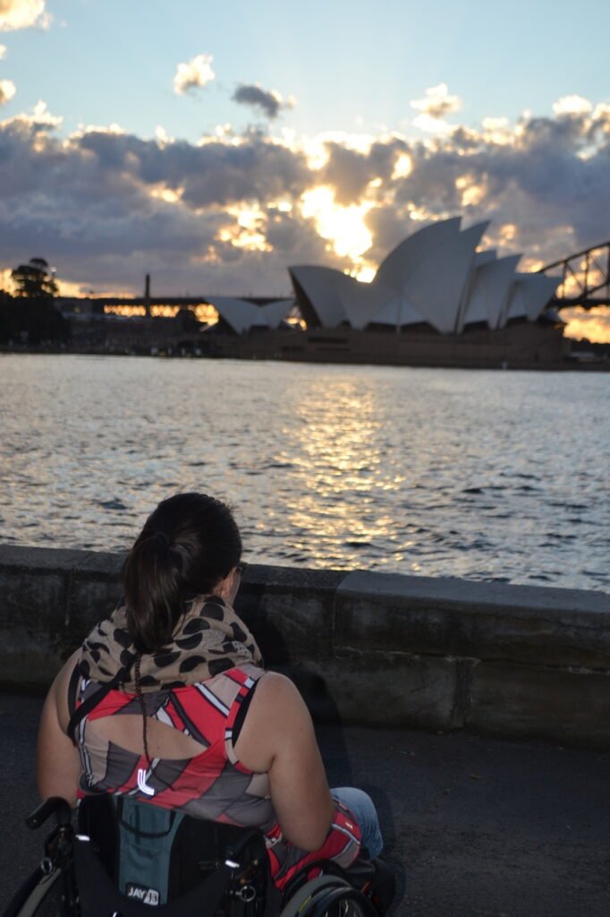 A woman in a wheelchair gazes at the Sydney Opera House, reflecting on the shimmering water at sunset.