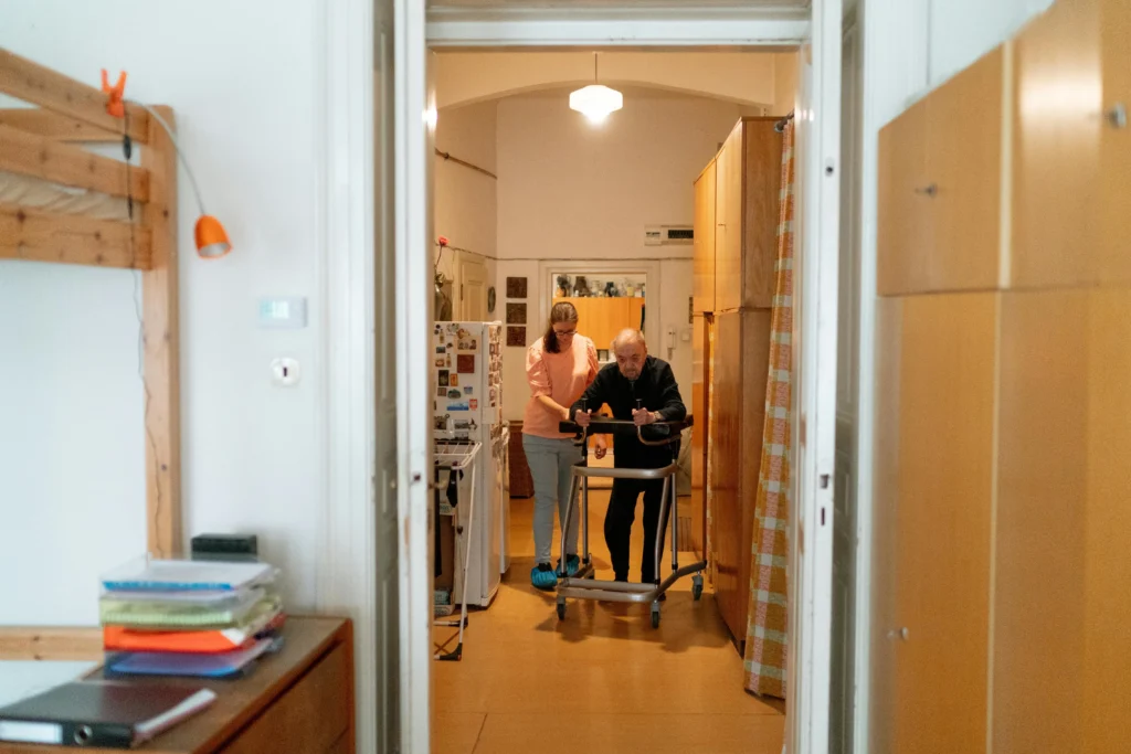 A woman helps an elderly person using a wheeled walker navigate a narrow hallway inside a home. The hallway is lined with wooden cabinets.