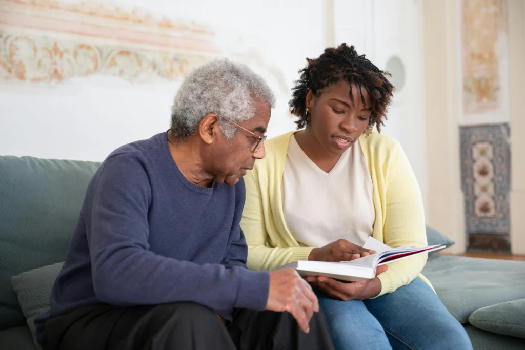 An elderly Black man with gray hair and glasses sits on a couch next to a younger Black woman with dreadlocks. They are both looking down at a book the woman is holding open.