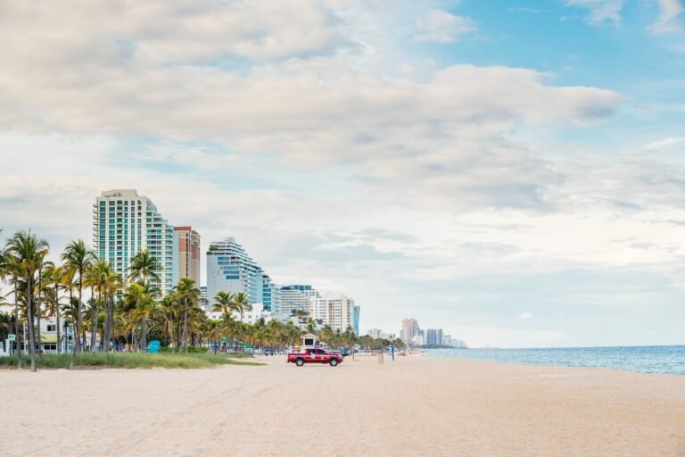 A beach scene featuring tall buildings, palm trees, and a red lifeguard truck on the sand under a cloudy sky.