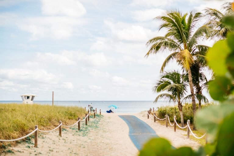 Sandy beach path lined with green grass and palm trees leads to the ocean. A blue umbrella and people are visible near the water, under a cloudy sky.