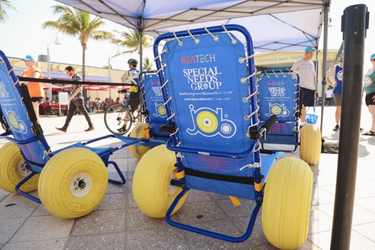 Beach wheelchairs with large yellow wheels and blue frames are displayed under a canopy. People walk and cycle nearby, palm trees in the background.