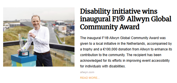 A man in a white collared shirt and blue jeans stands on a balcony and holds a large trophy. He is smiling slightly at the camera. In the background, there is a race track and other buildings under a cloudy sky.
