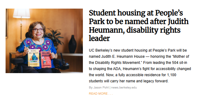 A woman in a wheelchair smiles at the camera while holding two books titled "Being Heumann" and "Rolling Warrior." She is in a room with a lamp and a dark vase in the background.