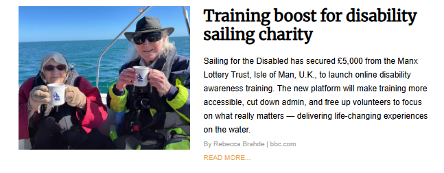 Two people are sitting on a sailboat holding mugs, smiling at the camera. The person on the left wears sunglasses and a red jacket, while the person on the right wears a hat, sunglasses, and a life vest over a blue jacket. The sea is visible behind them.