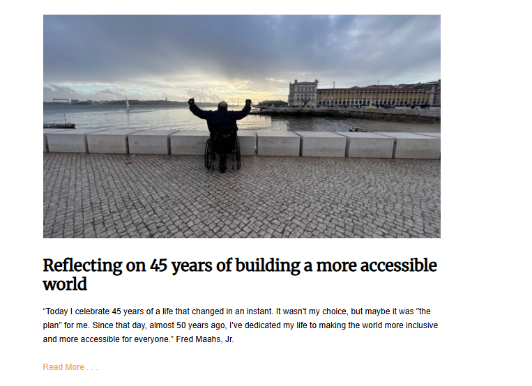 A person in a wheelchair sits with their back to the camera, arms raised in triumph. They are on a cobblestone path next to a stone barrier, with a body of water and buildings in the distance under a cloudy sky.
