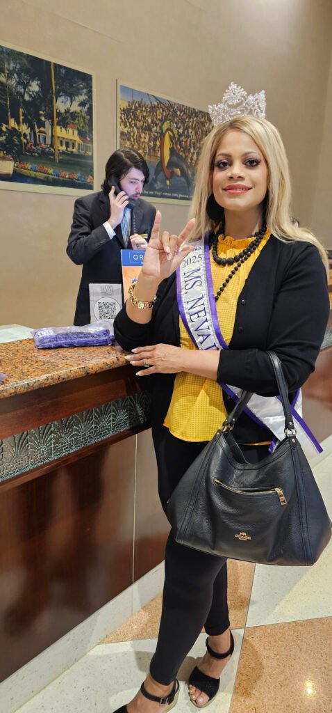 A woman wearing a crown and a "MS. NEVAD[sic] 2025" sash stands inside a building, posing for the camera and holding up a peace sign. She is wearing a yellow top, a black cardigan, and black leggings, and is holding a black Coach bag. A man is visible in the background talking on a phone at a counter.