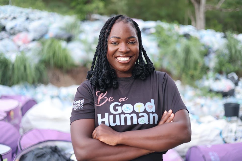 A close-up portrait of a smiling Black woman with her arms crossed, wearing a black t-shirt that reads "Be a Good Human" over a pile of trash and recycling bags in the background.