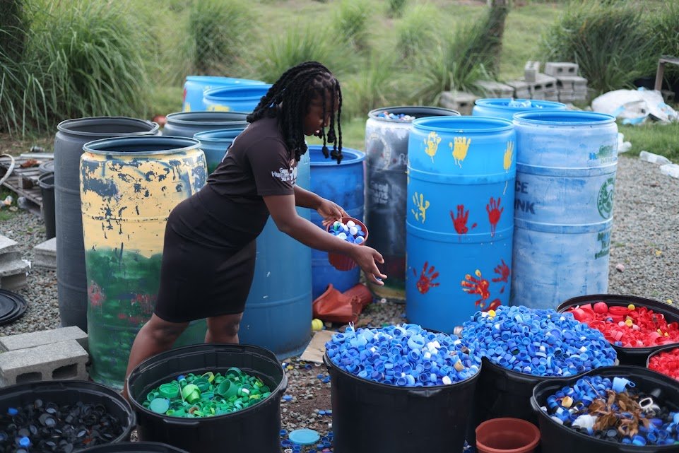 A Black woman in a black dress bends down to sort blue and white plastic bottle caps into a black bin amidst many other bins filled with various colored bottle caps (red, green, black) and several large, painted plastic barrels.