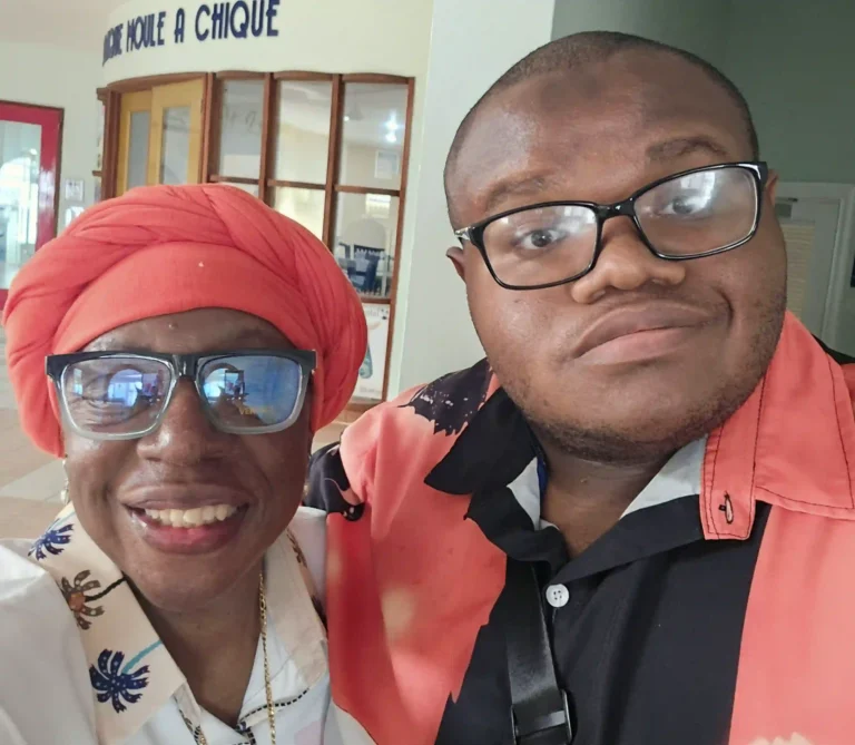 A smiling woman with a bright orange head wrap and dark sunglasses takes a selfie with a young man wearing glasses. They are standing indoors, and the man is wearing an orange and black patterned shirt.