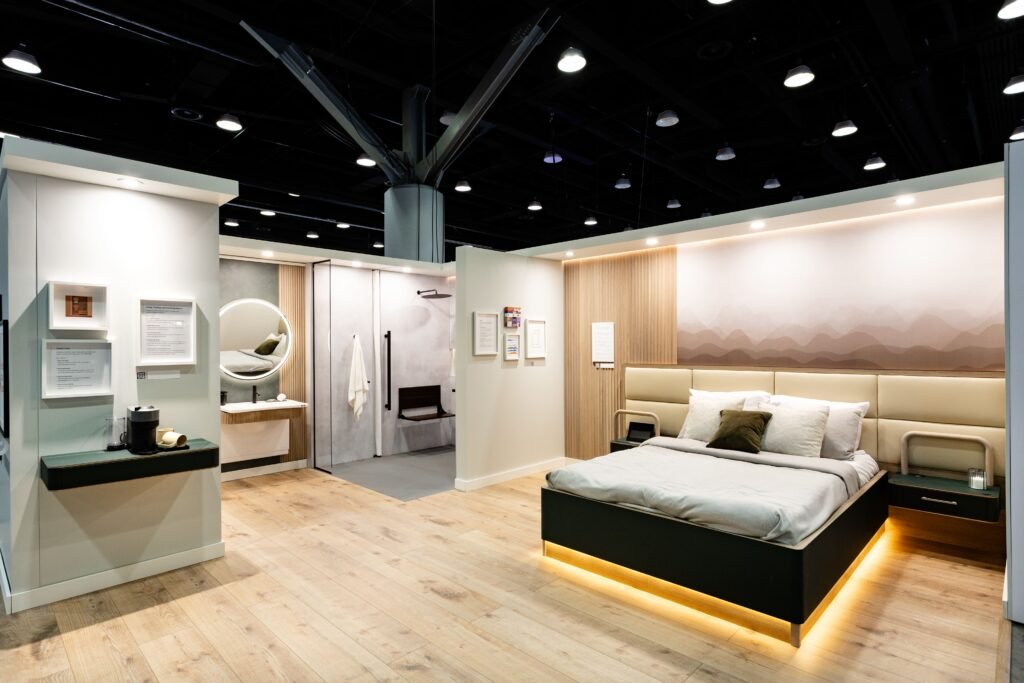 A wide, brightly lit view of an accessible design suite exhibit at a trade show. The suite features light wood flooring, a bed with a cream-colored upholstered headboard, and a low, dark frame with under-bed lighting. To the left, a separate area includes a modern bathroom with a round mirror and a bench in the shower area, showcasing accessibility features like grab bars. Informational signs are posted on the walls.