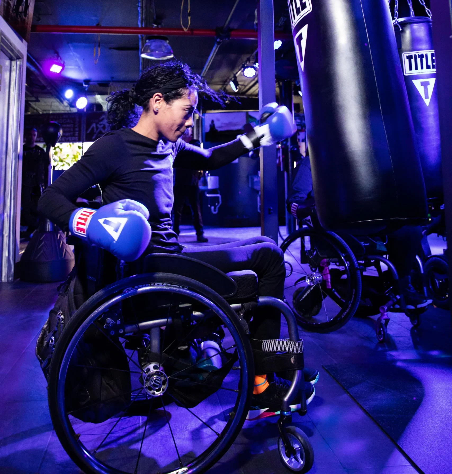 A woman with dark, curly hair in a wheelchair is boxing, throwing a punch with a blue glove toward a large black hanging punching bag that has the 'TITLE' logo in white. She is wearing a long-sleeved black top and black pants. The gym is dimly lit with vibrant purple and blue lighting. Another wheelchair is visible to the right.