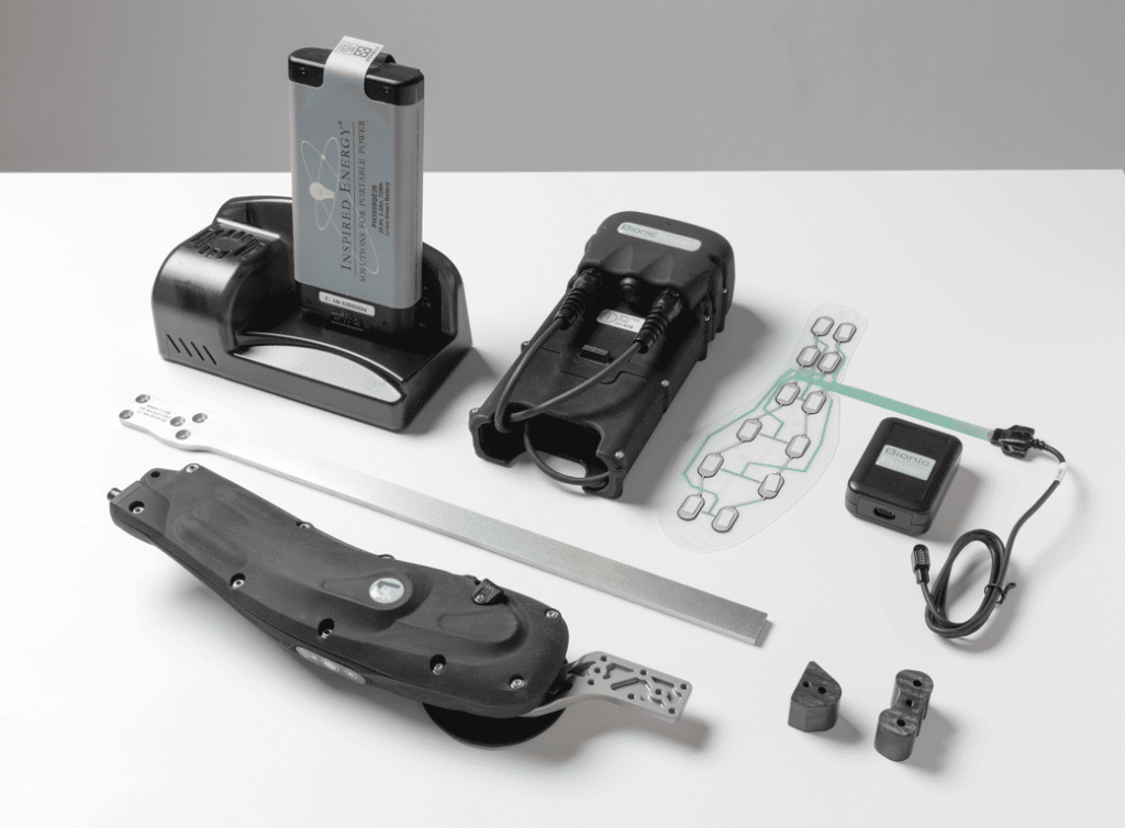 A flat-lay image showing the complete set of components for the Bionic Power Agilik energy-generating system. Key parts include a large, metallic battery pack labeled "Inspired Energy" standing in its charging base, a central electronic control unit with two cables attached, and a long, curved energy harvesting unit designed to attach to a prosthetic or orthotic device. Other components visible are a flexible insole-like sensor pad with a circuit tracing, a small black power adapter, and two small black joint pieces. The parts are arranged on a clean white surface.