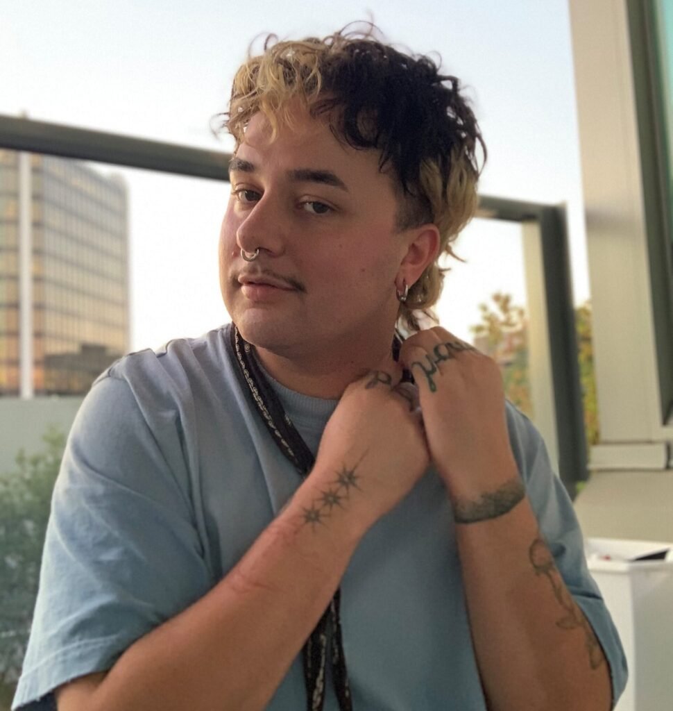 A portrait of Bug Cru (a non-binary person) standing near a large window with a cityscape visible in the background. They have a dark, curly mullet with blonde highlights at the front and are wearing a light blue t-shirt with a braided cord necklace. They have multiple tattoos visible on their hands and arms, including small stars and text, and are touching their hands together near their chest while looking directly at the camera.