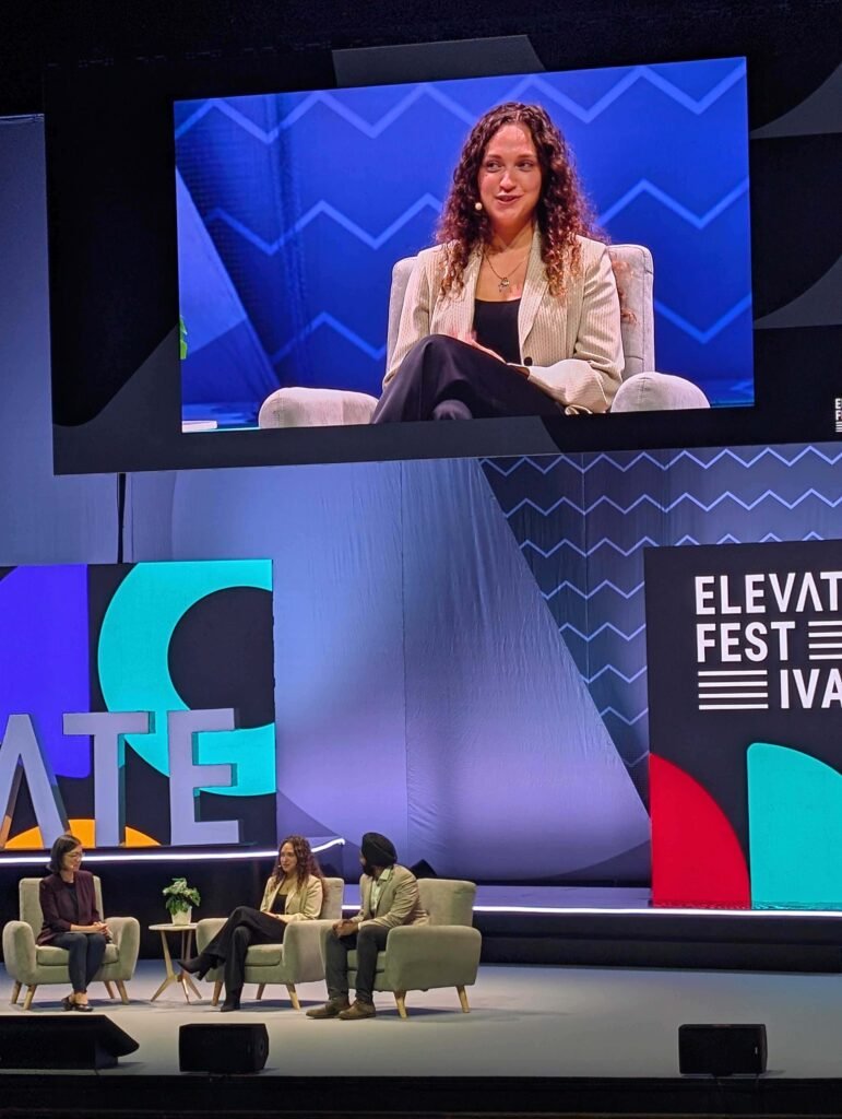 photograph of a panel discussion at an event labeled ELEVATE FESTIVAL. Three panelists are seated in light grey armchairs on a stage. A large screen above the stage shows a close-up of one of the female panelists, who has long curly hair and is wearing a striped blazer. The stage features bold, colorful graphic signage reading "ELEVATE."