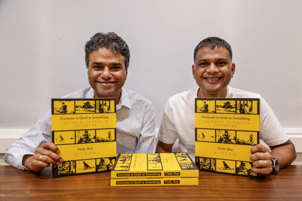 Two men are seated at a table, both smiling brightly as they hold up copies of the book "Everyone is Good at Something" by Vicky Roy. The book has a yellow cover with black and white images and text, including a section in braille. Several more copies are stacked on the table in front of them.