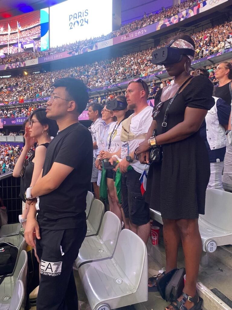 A diverse group of spectators standing in the seating area of a large stadium, likely at the Paris 2024 Olympic Games, based on the overhead screen graphic. Several individuals in the group, including a woman in the foreground and a man behind her, are wearing GiveVision smart headsets (or similar visual aids) to watch the event. The stadium is packed with people.