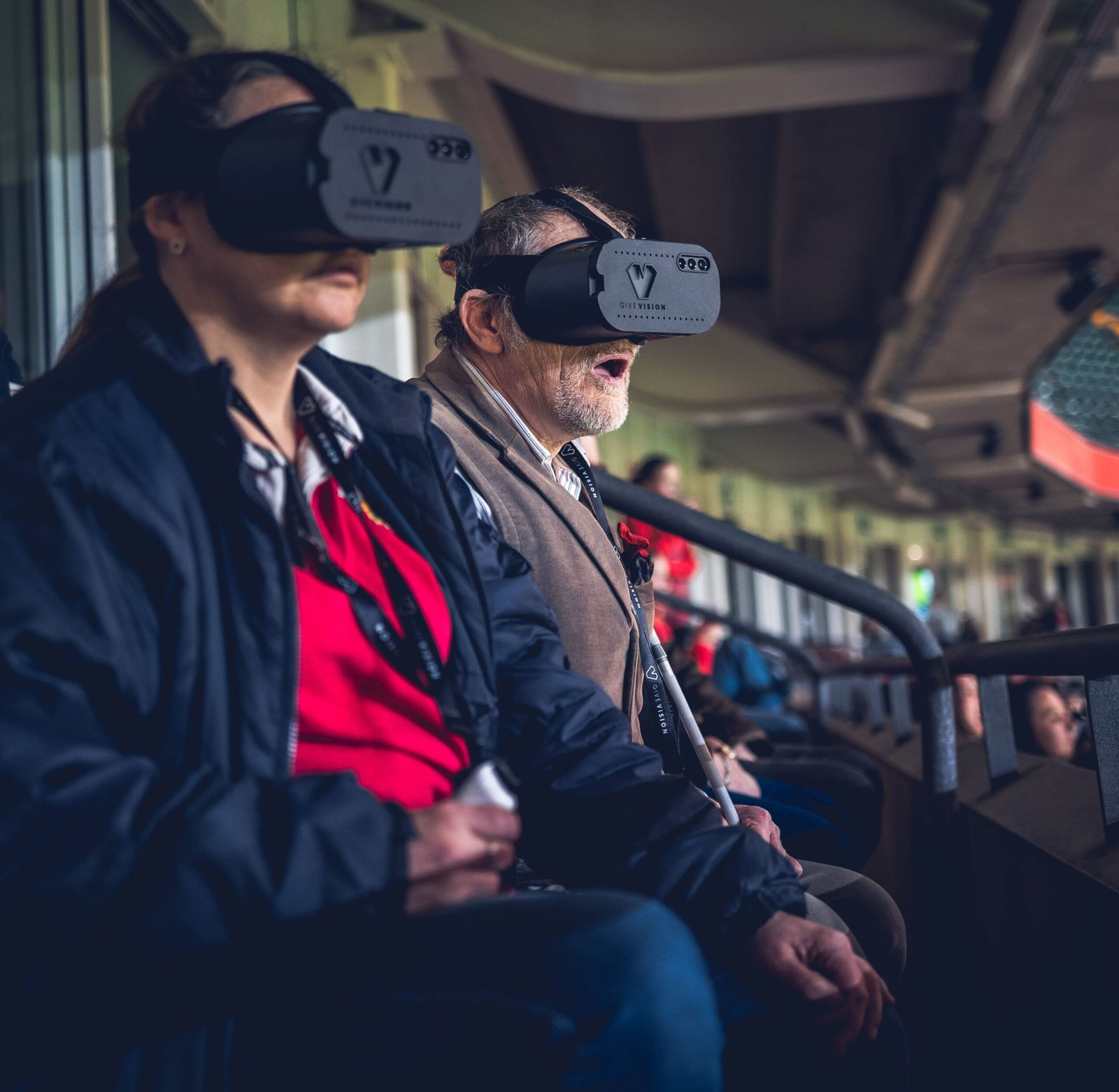 Two spectators, a woman and an older man, are seated side-by-side in a stadium wearing GiveVision smart headsets (or similar visual aids) over their eyes. The man appears to be reacting enthusiastically to the event, while the woman looks focused. The scene is dimly lit inside the stadium seating area, with a railing in the foreground.