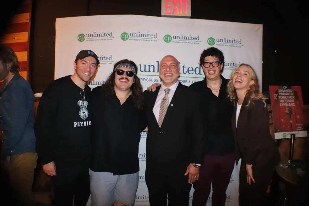 A group of five people—four men and one woman—are posing together and smiling in front of a white backdrop covered with a repeating green logo for 'cp unlimited PROGRESS WITH PURPOSE & CARE'. The center man is wearing a suit and tie, while the others are dressed more casually, including one man on the left wearing a 'PHYSICS' sweatshirt and another with a bandana and sunglasses.
