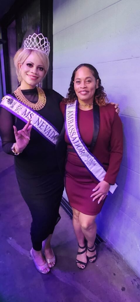 A full-length photo of two women standing side-by-side against a purple-lit wall. The woman on the left is wearing a crown, a black dress, and a sash that reads "Ms. Nevada 2024," and is making a peace sign. The woman on the right is wearing a maroon dress/blazer and a sash that reads "Ambassador Nevada 2025."