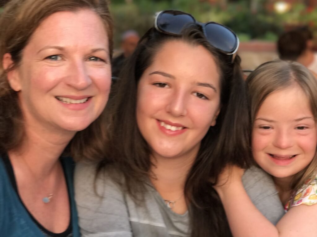 A close-up selfie of a smiling woman and two young girls, possibly taken at an outdoor restaurant. The woman is on the left, and the girl on the right has Down syndrome. They are all looking directly at the camera.