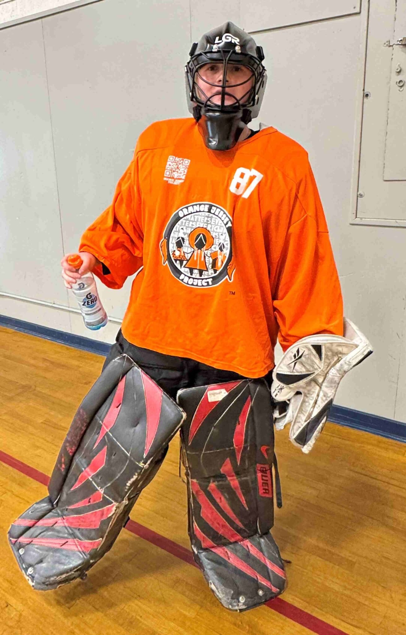 Indoor hockey goalie wearing orange Orange Series Project jersey number 87, black and red Bauer goalie pads, black LGR helmet, and white glove, holding a Gatorade Zero bottle on a gym floor