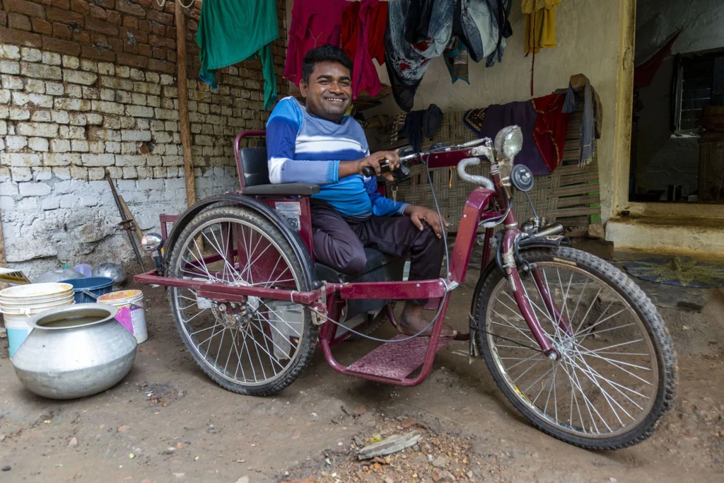 A man named Jagmohan Thakur with short stature and a bright smile sits on a custom-made, hand-operated, three-wheeled vehicle, which resembles a motorized tricycle or cycle-rickshaw. He is wearing a blue and white striped long-sleeve shirt and dark pants. He is indoors in a rustic area with a brick wall behind him and clothing hanging to dry overhead. A large metal pot sits on the dirt floor to the left.