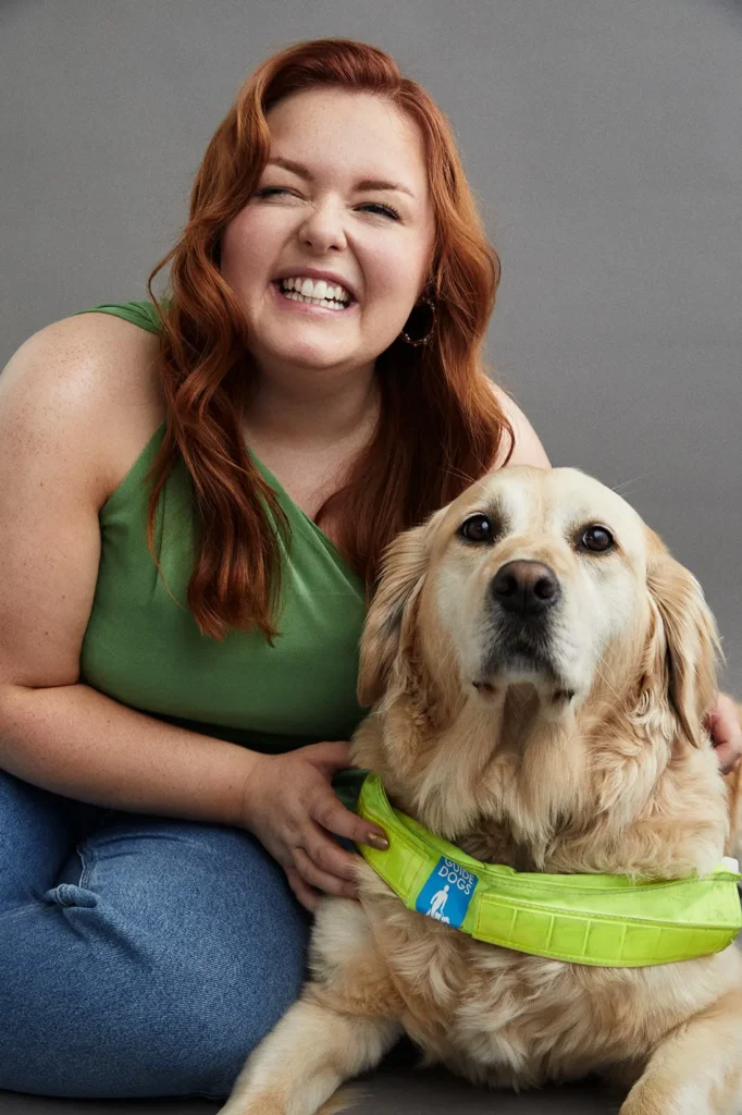 A portrait of a young woman with long, wavy red hair smiling widely while sitting next to a golden retriever guide dog. The woman is wearing a green sleeveless top and blue jeans, with her arm wrapped around the dog. The dog is looking directly at the camera and wearing a reflective, bright yellow guide dog harness. The background is a solid medium gray.