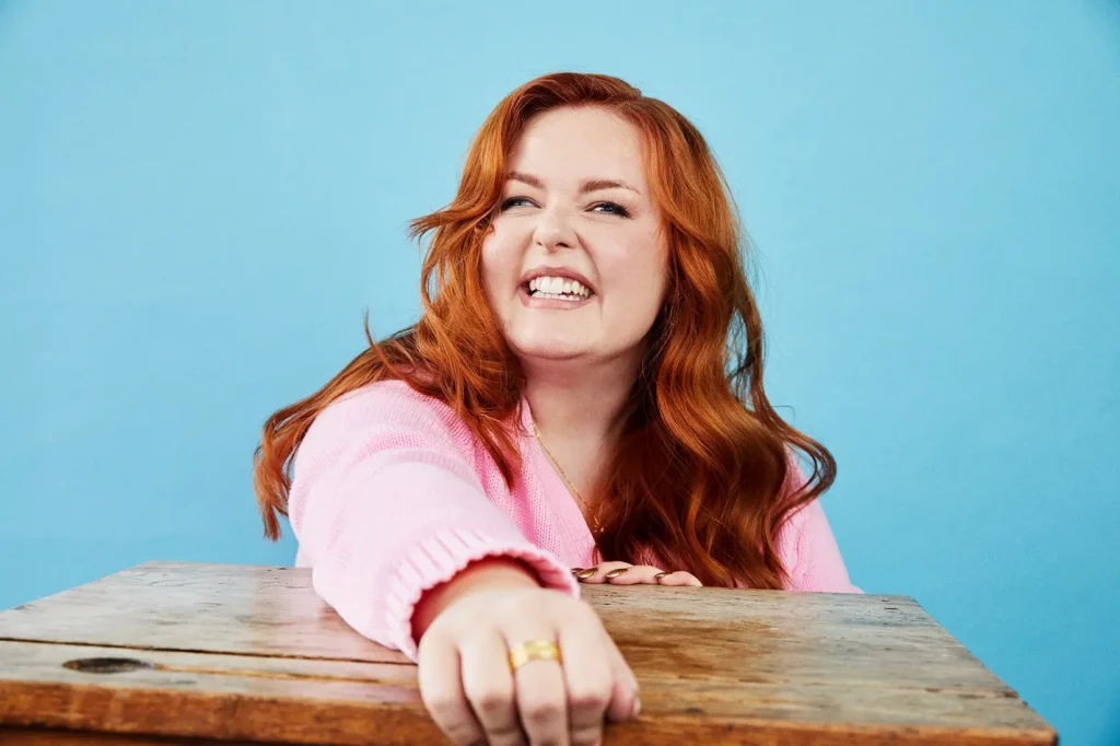 A close-up portrait of a young woman with long, flowing red hair, leaning forward over a wooden desk or table and smiling widely, showing her teeth. She is wearing a light pink, ribbed, long-sleeved top and a gold ring. The background is a solid bright blue.