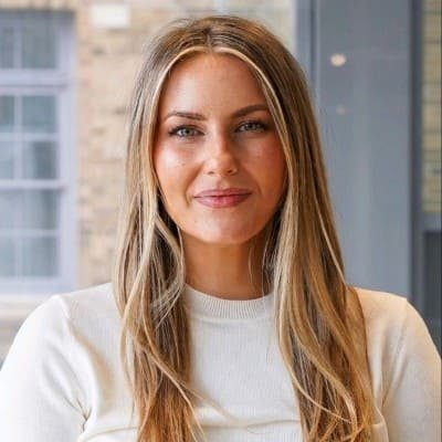 A brightly lit professional headshot of Morgan Lorimer. She is a young woman with long, straight blonde hair with lighter highlights framing her face, and she is wearing a white crewneck top. She is looking directly at the camera with a slight smile. The background is softly blurred, showing an interior setting with a window.