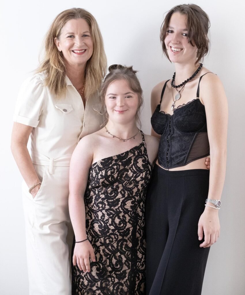 A studio portrait of three smiling females standing against a white background. A woman on the left wears a cream-colored jumpsuit. A young woman in the center, who has Down syndrome, wears a black, one-shoulder dress with a lace overlay. A young woman on the right wears a black corset top, black pants, and a thick black necklace.