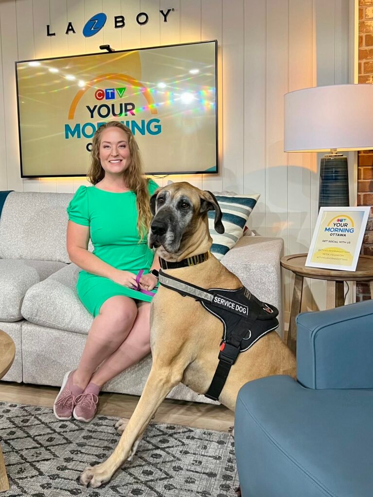 A photo of Michelle Weger and her service dog, Quinn, on the set of CTV's Your Morning show. Michelle is seated on a couch, smiling and wearing a bright green dress and pink running shoes. Quinn, the large Great Dane service dog wearing a black harness, is seated politely next to her. A monitor in the background displays the show's logo, and the set features comfortable furniture and a patterned rug.