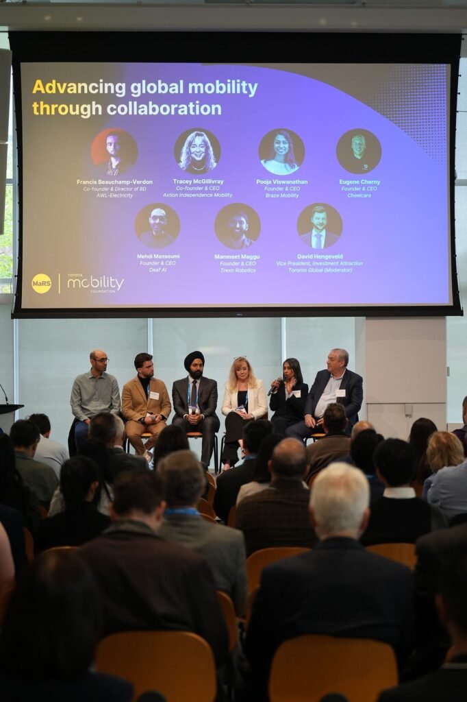 A wide shot of a panel discussion titled "Advancing global mobility through collaboration" taking place at the MaRS Toyota Mobility Forum. Seven panelists are seated on stage, facing a large, engaged audience. A screen above them displays their names and titles, including Tracey McGillivray of Axtion Independence Mobility, Pooja Viswanathan of Braze Mobility, and Manmeet Maggu of Trexo Robotics. The moderator, David Hengeveld, is seated on the far right.