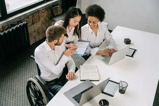 Three diverse colleagues collaborating at a white office table, including a man using a wheelchair, looking at a shared notebook.