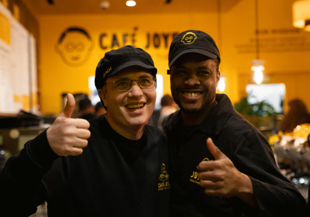 Two men, both wearing black shirts and black caps, stand together smiling brightly and giving thumbs up gestures in what appears to be a cafe. The man on the left wears glasses. The background is a bright yellow wall with the words "CAFÉ JOYEUX" and a stylized face logo written on it in black.