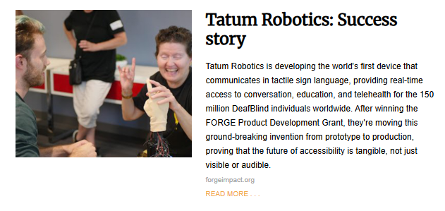 A candid photo accompanying the article "Tatum Robotics: Success story." A smiling woman who is DeafBlind is seated at a table, touching a realistic, prosthetic hand prototype that is wired with electronics. She appears to be demonstrating or testing a device that provides tactile sign language. A man with a beard and light green hair is sitting across from her, watching. The background is a blurred image of an office or workshop.
