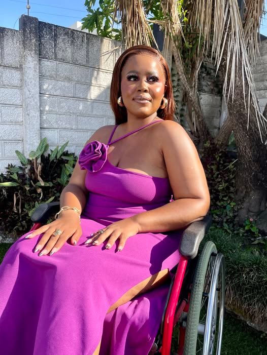 Thembelihle Ngcai sits in a red and black wheelchair in a sunny outdoor setting. She is wearing a striking, bright purple one-shoulder gown with a fabric rose detail on the strap, complemented by gold jewelry. She has a warm expression and is looking slightly off-camera to the right, with a light brick wall and lush greenery behind her.