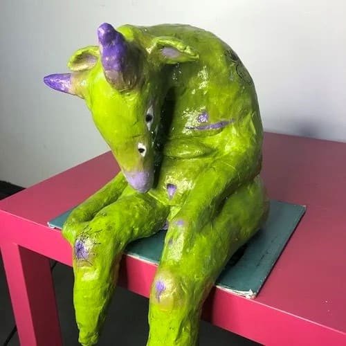 A photograph of an untitled sculpture. The figure is a small, seated, lime-green creature with horns, appearing to be a bovine or mythological animal, created from a textured material like papier-mâché or clay. It has purple accents on its horns, knees, and body, and is slumped over, looking downward in a mournful or pensive pose. The sculpture is sitting on a small, dark object placed on a brightly pink table.