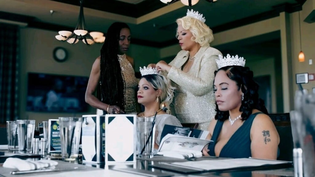 Four women seated at a formal table in a restaurant. A blonde woman in a sparkling gold blazer is placing a crown on the head of another woman seated to her left. Two other women, one standing and one seated, are also visible.