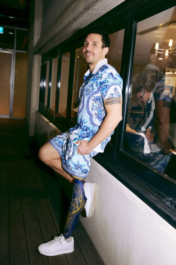 A man with dark hair and a small beard is leaning casually against a dark window frame outdoors at night. He is wearing a matching short-sleeved button-down shirt and shorts set with an intricate blue and white baroque-style pattern. He is wearing a dark blue compression sock or legging with a bright gold graphic design on his right leg, and white sneakers. His right hand is in his shorts pocket, and he is looking directly at the camera with a slight smile. A faint reflection of two people is visible in the window behind him.