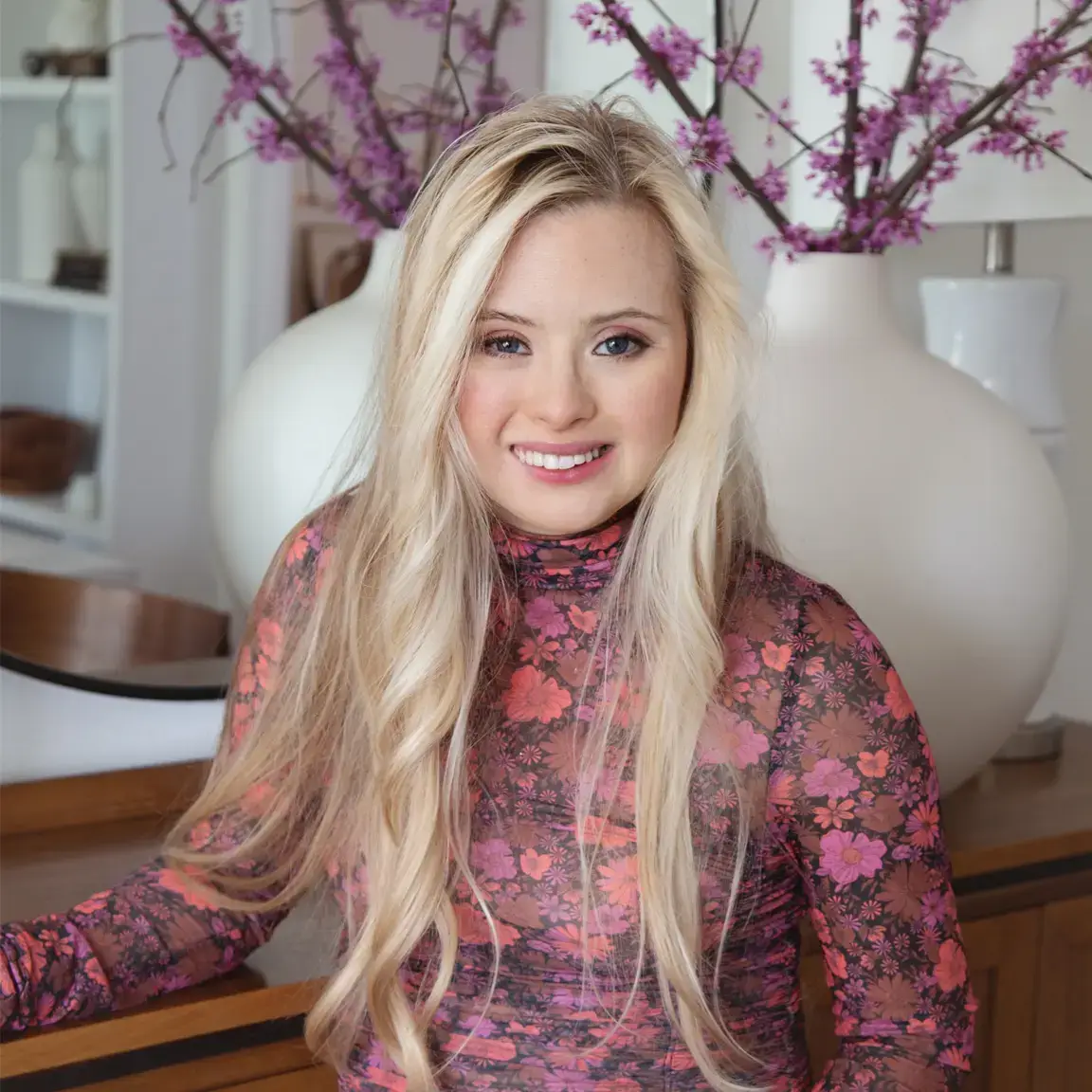 A close-up portrait of a smiling blonde woman with Down syndrome, wearing a sheer floral pink and orange turtleneck top, standing in front of large white vases and pink flowering branches.