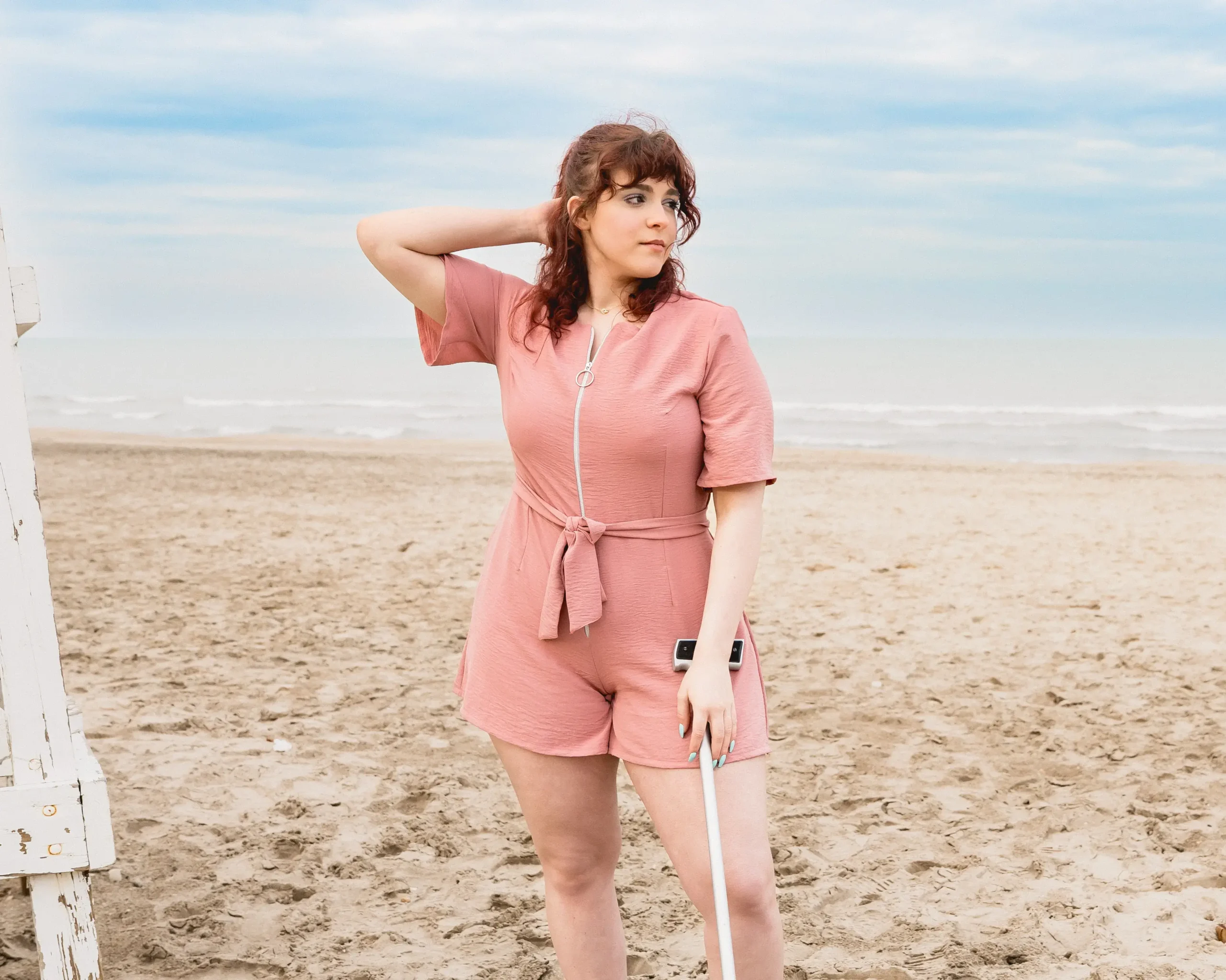 A woman with curly reddish-brown hair stands on a beach, looking toward the right. She is wearing a mauve pink adaptive romper with short sleeves, a front zipper, and a tied waist. She holds a white cane in her left hand and touches the back of her head with her right hand. The ocean and sky are visible in the background.