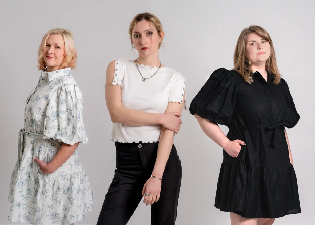 A professional studio portrait of three women standing side-by-side against a plain white background. On the left, a blonde woman smiles while wearing a white and light-blue floral dress with puff sleeves. In the center, a blonde woman with a serious expression wears a white ribbed top and black trousers. On the right, a woman with light brown hair wears a black dress with voluminous puff sleeves and a ruffled hem.