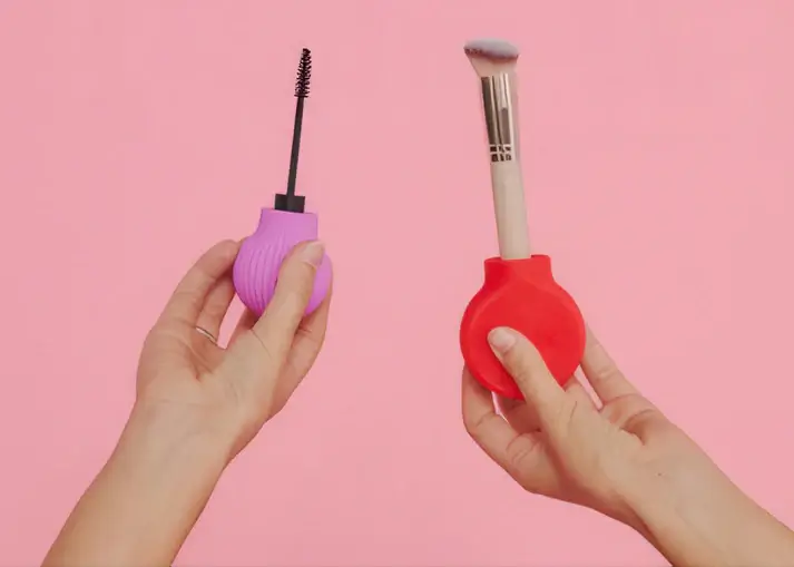 A close-up shot against a pink background shows two hands holding makeup tools. The hand on the left holds a mascara wand with a purple, ribbed, bulb-shaped silicone handle. The hand on the right holds a foundation or powder brush with a wooden handle inserted into a red, smooth, bulb-shaped silicone grip.