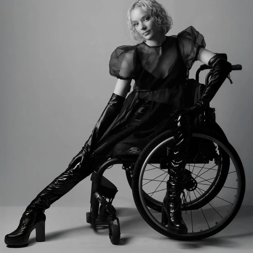 Black-and-white fashion portrait of a woman seated in a wheelchair, wearing a sheer black dress with puffed sleeves, long black gloves, and high platform boots. She poses confidently against a neutral studio background
