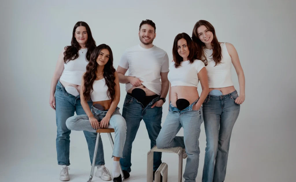 A group of five smiling individuals—three women and two men—stand and sit together in a bright, white studio. All are wearing white t-shirts or tank tops and denim jeans, with their waistbands partially pulled down to expose their lower abdomens, where ostomy pouch covers of various colors (beige and black) are visible.