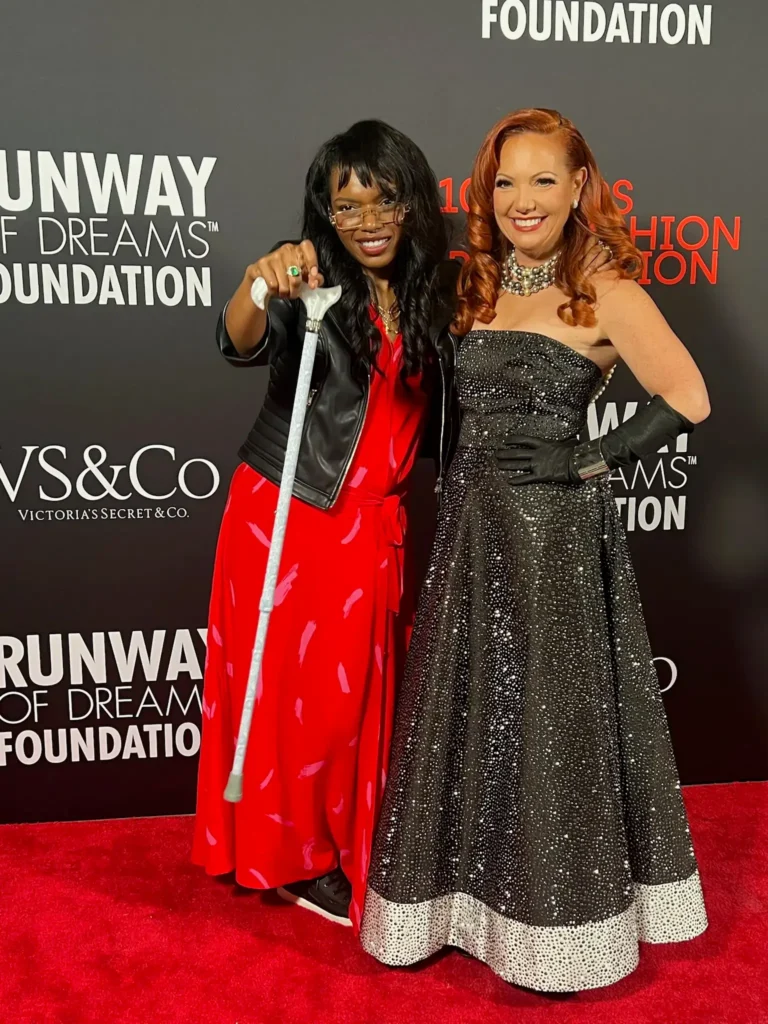 Two women posing together on a red carpet. On the left, a woman wearing glasses, a leather jacket, and a red dress holds a white mobility cane up playfully. On the right, a woman with red hair in a black formal gown smiles with her arm around her companion. The background displays the text 'Runway of Dreams Foundation'.