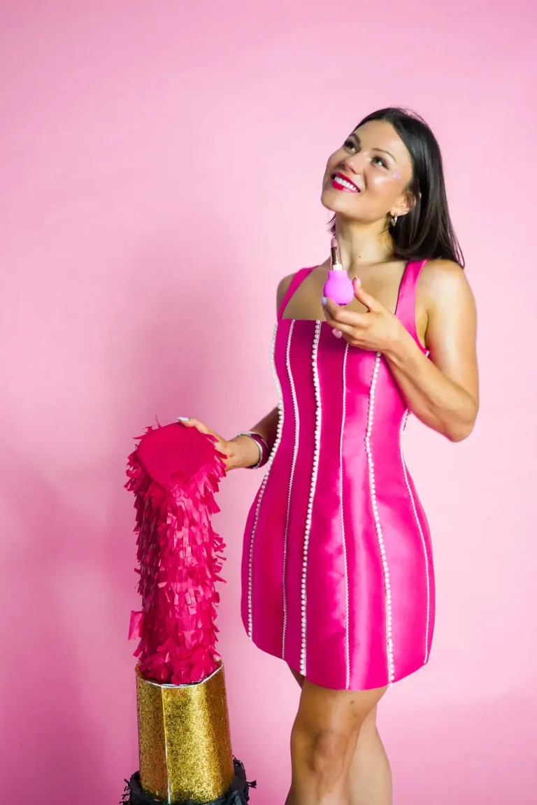 A woman in a vibrant, pink minidress with vertical pearl-like beading, smiling and looking up against a bright pink background. She holds a small pink, bulbous-shaped adaptive grip attached to a lipstick tube. Next to her is an oversized novelty lipstick prop with a golden base and pink fringed top.