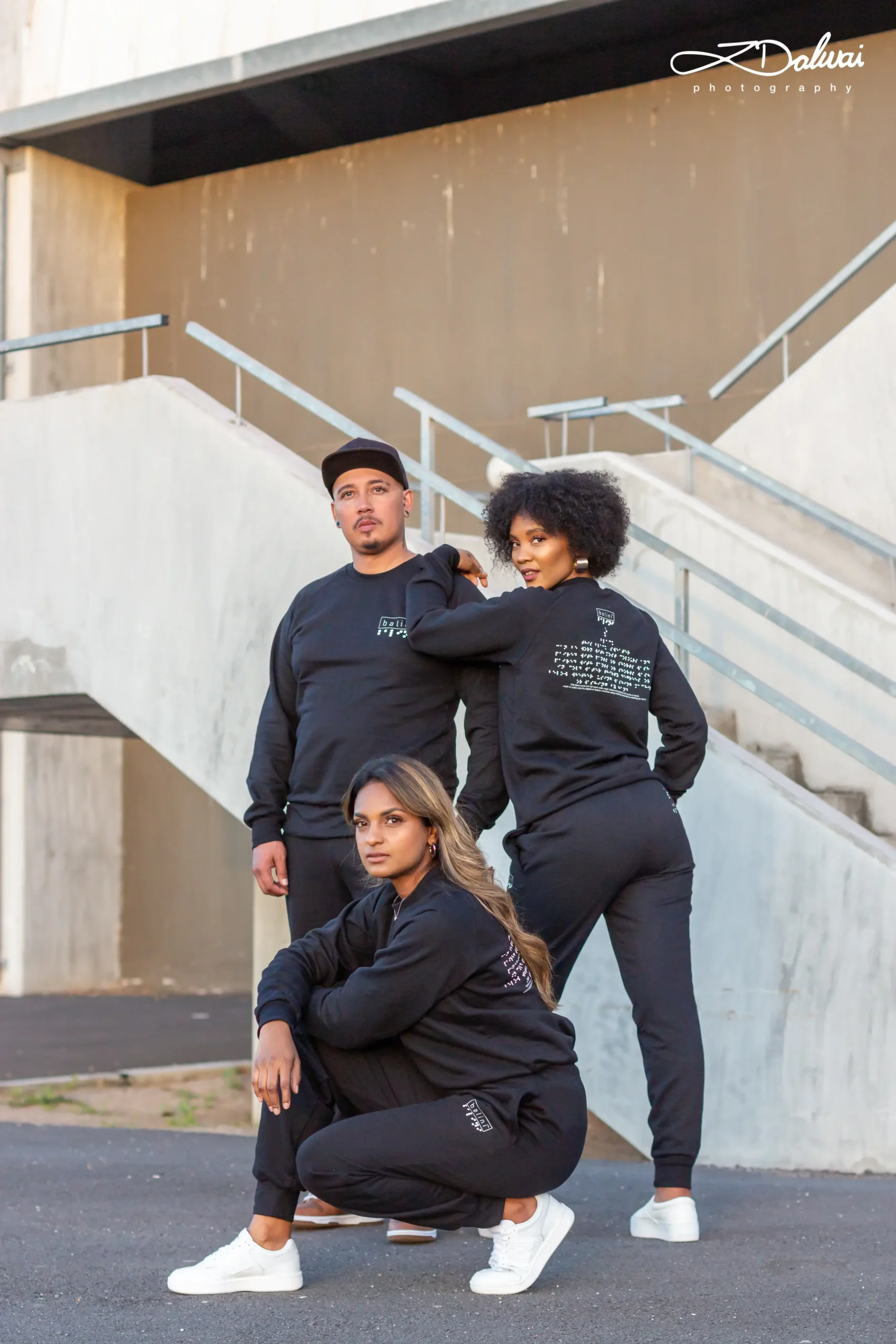 A vertical split image against a white background featuring two models walking toward the viewer. On the left, a man walks wearing a light tan bomber jacket over a white collared shirt and light khaki trousers, paired with white sneakers. On the right, a woman stands with her legs crossed, wearing a white sleeveless crop top and a pleated, two-toned skirt in tan and light pink, paired with open-toe nude heels.